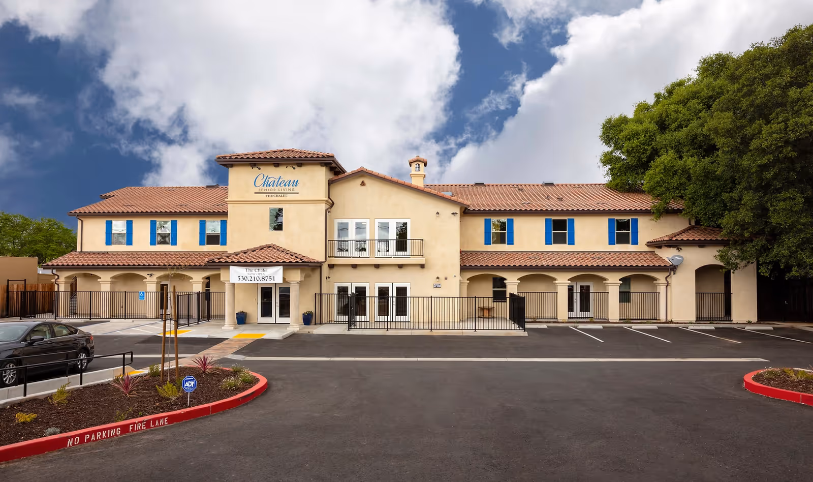 Front exterior of a two-story Mediterranean-style senior living building called The Chalet with a parking lot and landscaped curb in front.