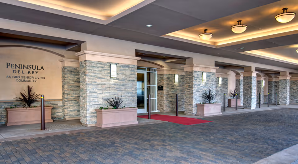 Entrance area of Peninsula Del Rey senior living community featuring stone pillars, a covered driveway with a red carpet, potted plants, and ceiling lights.