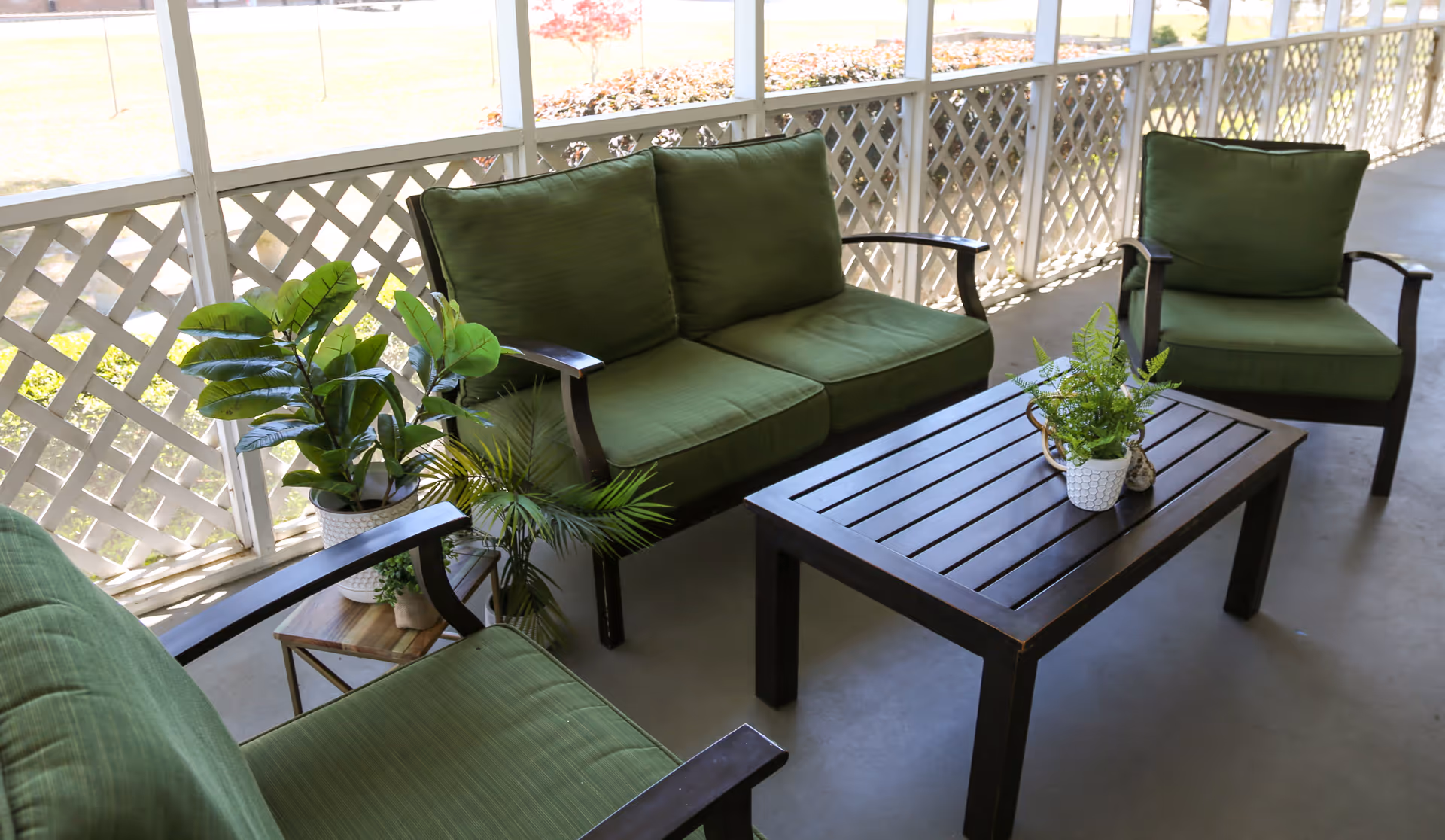 Screened porch seating area with green-cushioned patio chairs and a dark wooden slatted coffee table topped with potted plants.