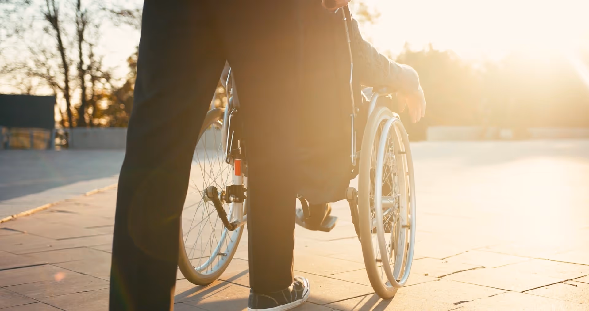 Back of a person pushing a wheelchair on a sunlit outdoor plaza.