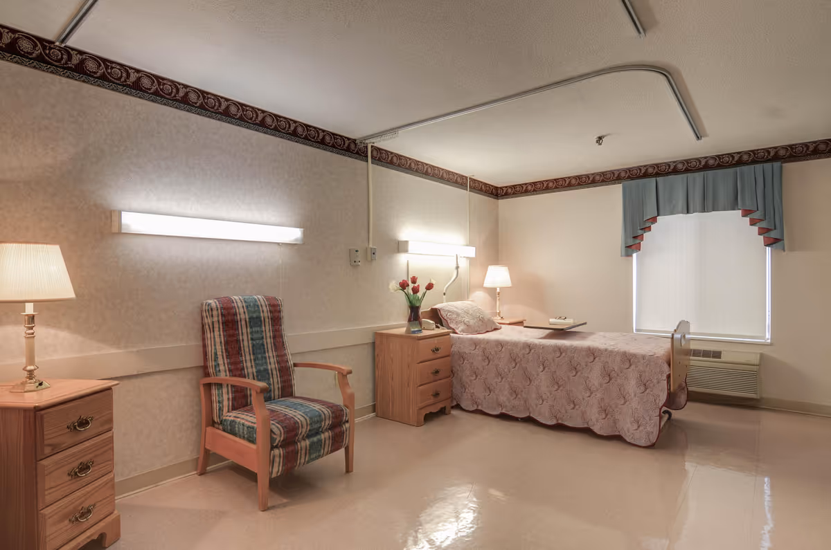A simple, clean senior living bedroom with a single bed covered in a pink patterned bedspread, a wooden nightstand with a vase of flowers and a lamp, a striped upholstered armchair, and a window with blue and red valance curtains. The room has beige walls with a decorative border near the ceiling and polished tile flooring.