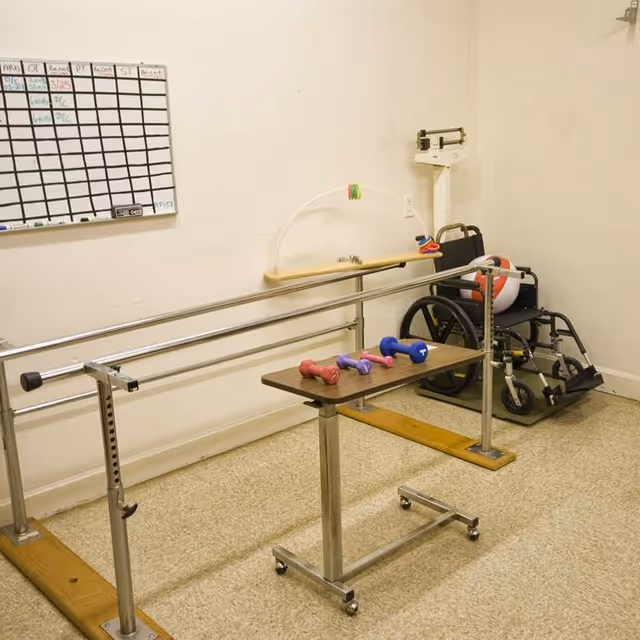 Therapy room in a facility with parallel bars, a wheelchair, and weights.