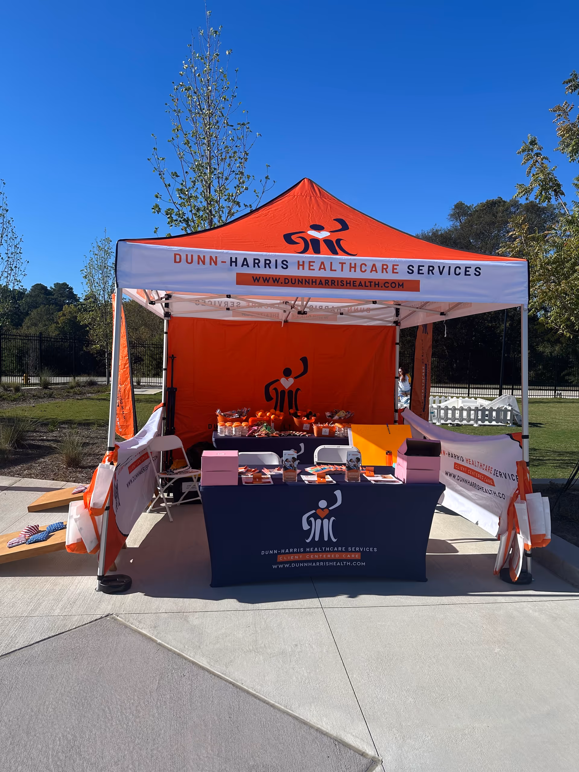 Outdoor Dunn-Harris Healthcare Services tent with tables and promotional materials in a sunny park.