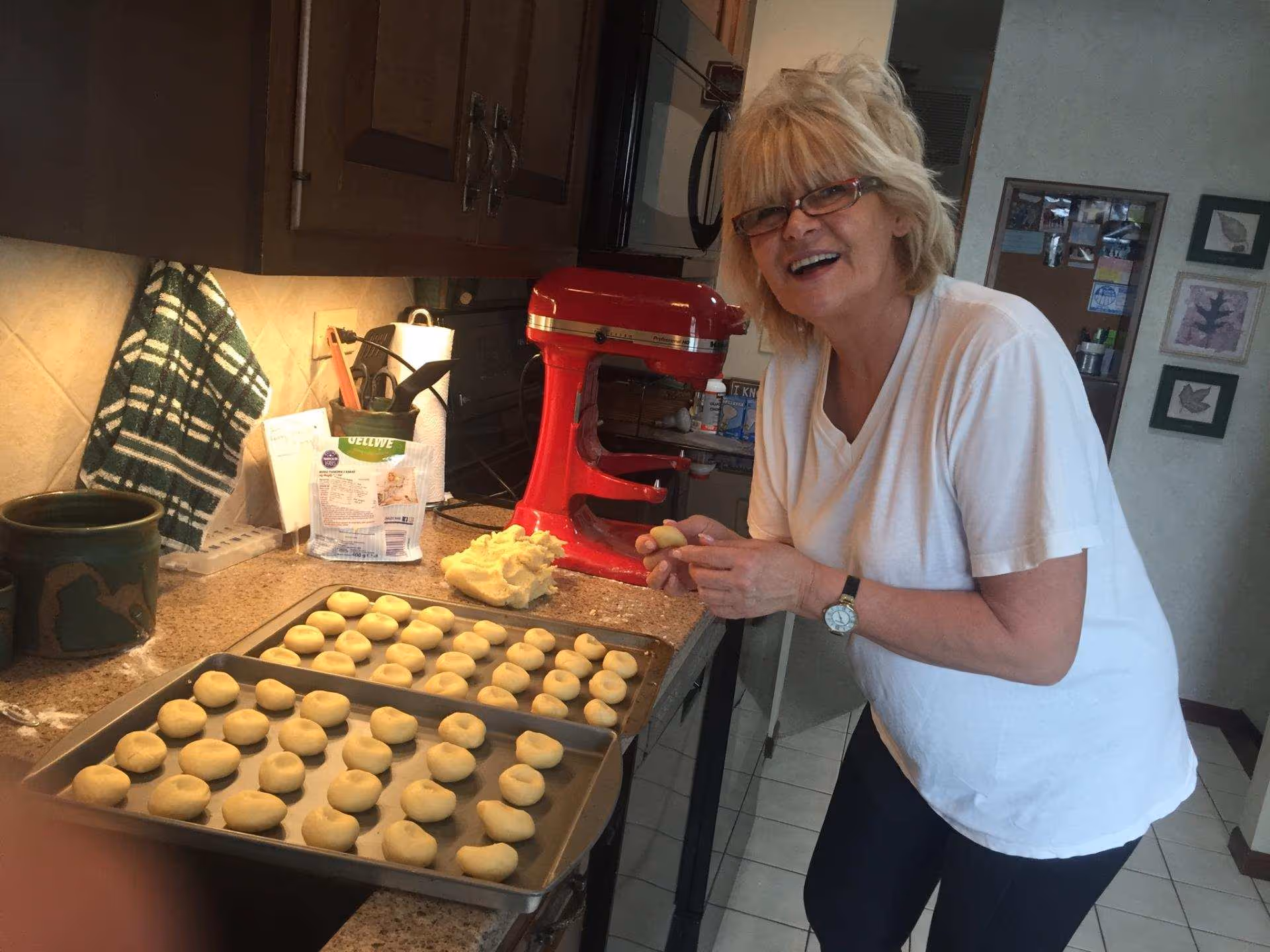 Smiling woman in a white shirt prepares dough beside a red mixer in a kitchen.