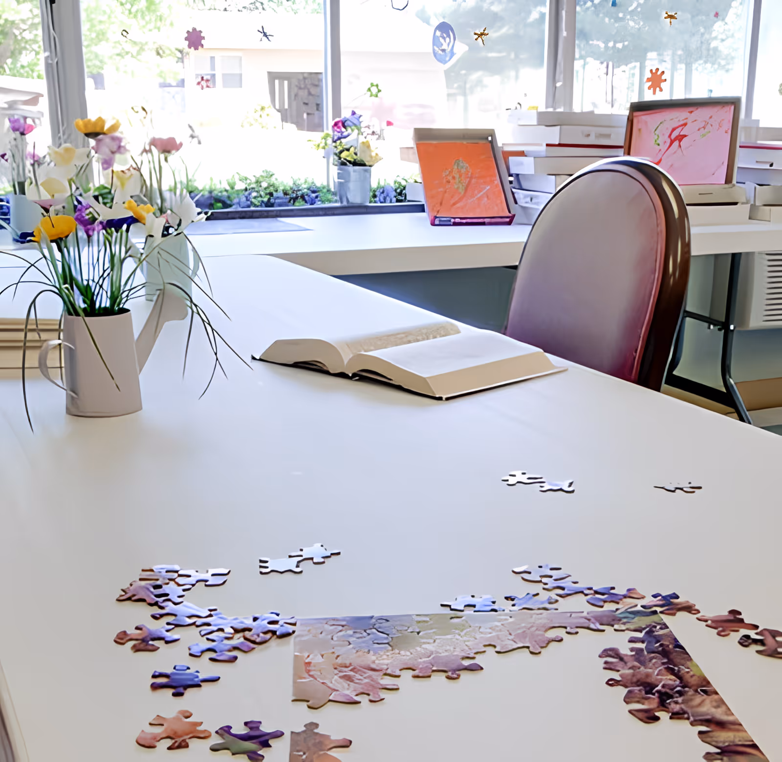 A bright room with a large white table featuring an open book, scattered puzzle pieces, and a white watering can with colorful artificial flowers. In the background, there are windows with decorative stickers and shelves with stacked boxes and framed artwork.