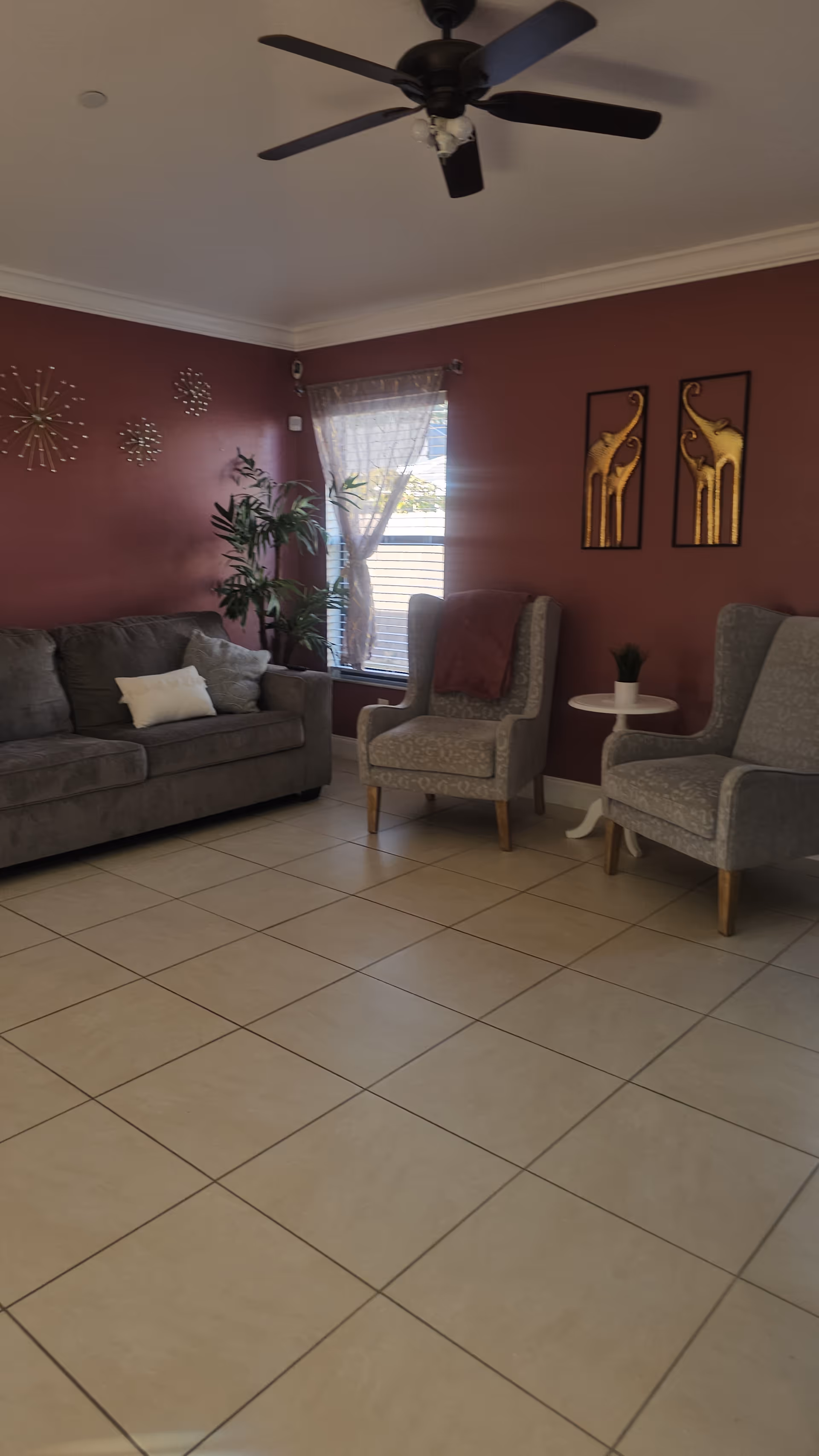 Living room in an assisted living facility with maroon walls, seating, plant, and ceiling fan.