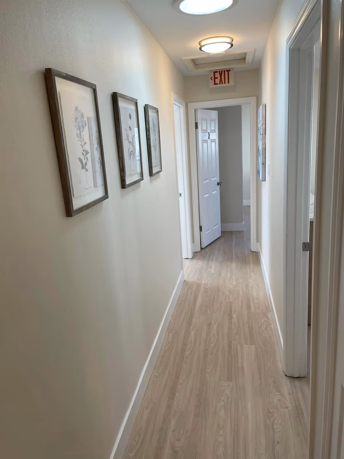Long beige corridor in a senior living facility with framed botanical art and open doors.