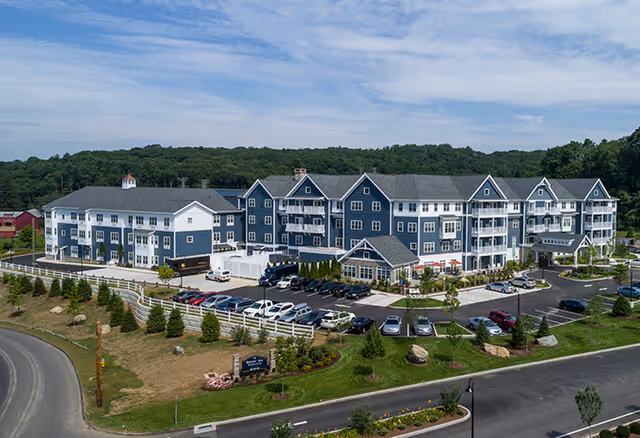 A large, multi-story senior living facility building painted in blue and white with multiple balconies and a parking lot filled with cars in front. The building is surrounded by landscaped greenery and trees, with a road curving around the property. The sky is partly cloudy.