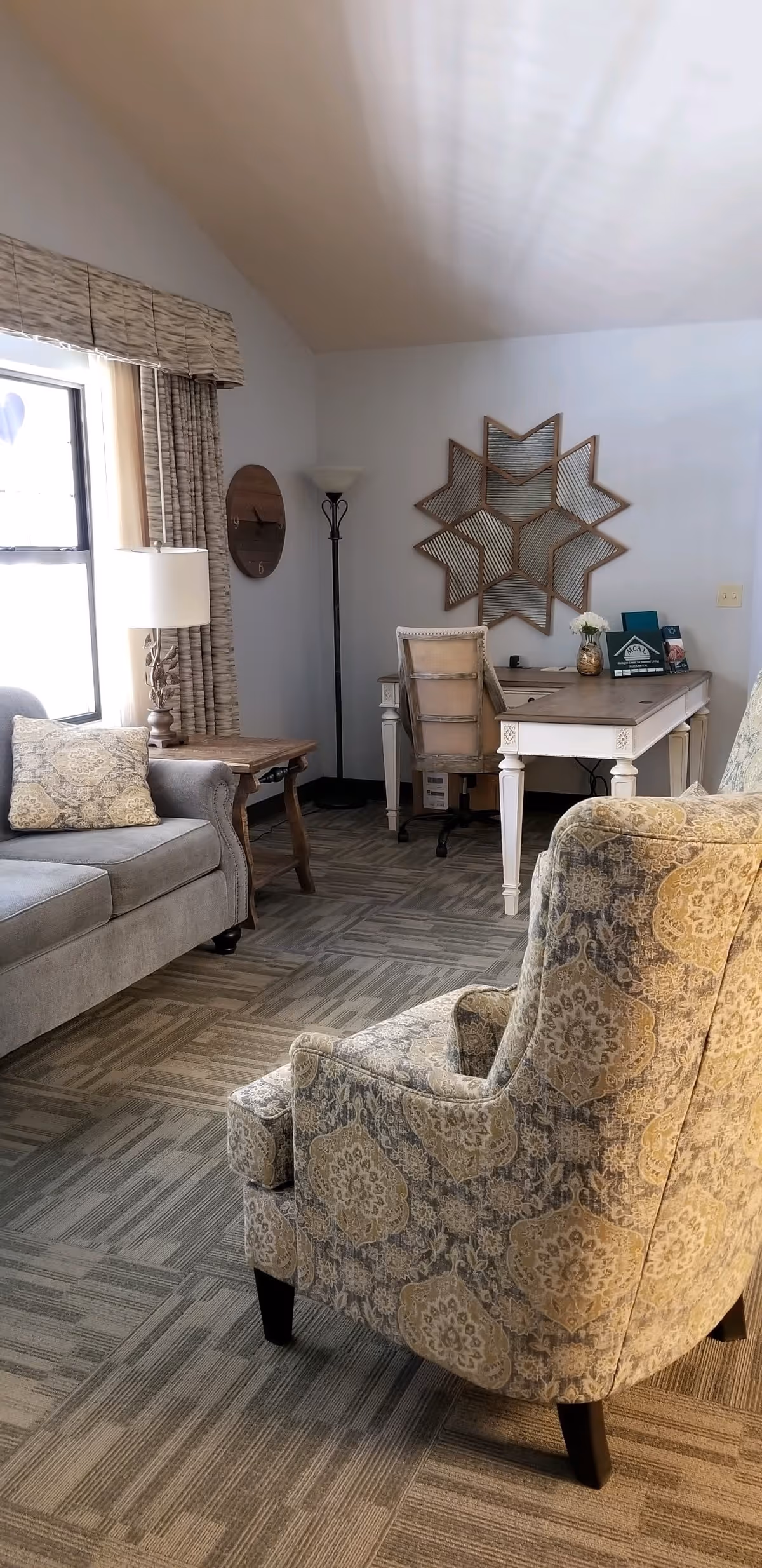 A cozy living room area with a patterned armchair in the foreground, a gray sofa with a decorative pillow near a window with curtains, a wooden side table with a lamp, and a white desk with a chair against the wall. A decorative star-shaped mirror and a floor lamp are also visible.
