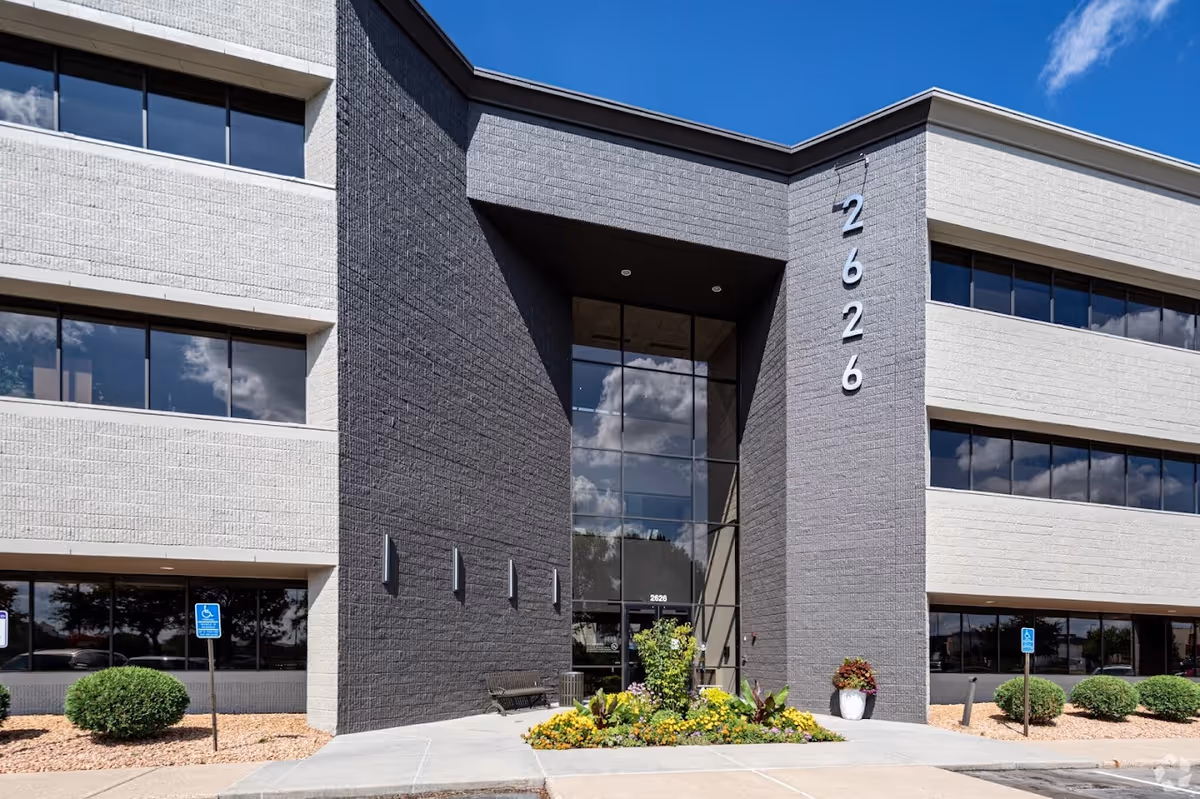 Modern two-story building entrance with glass doors, gray and white facade, and landscaped front.
