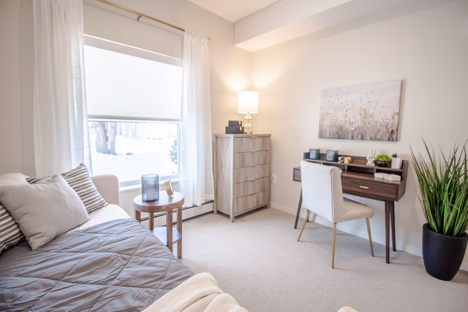 Bright bedroom with a bed, desk, dresser, and large window in a neutral room.