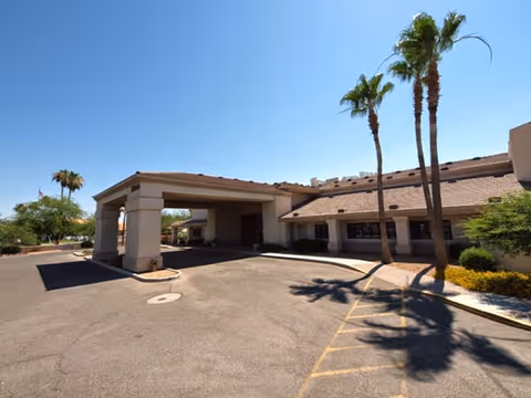 Exterior view of a single-story building with a covered entrance driveway, surrounded by palm trees and desert landscaping under a clear blue sky.