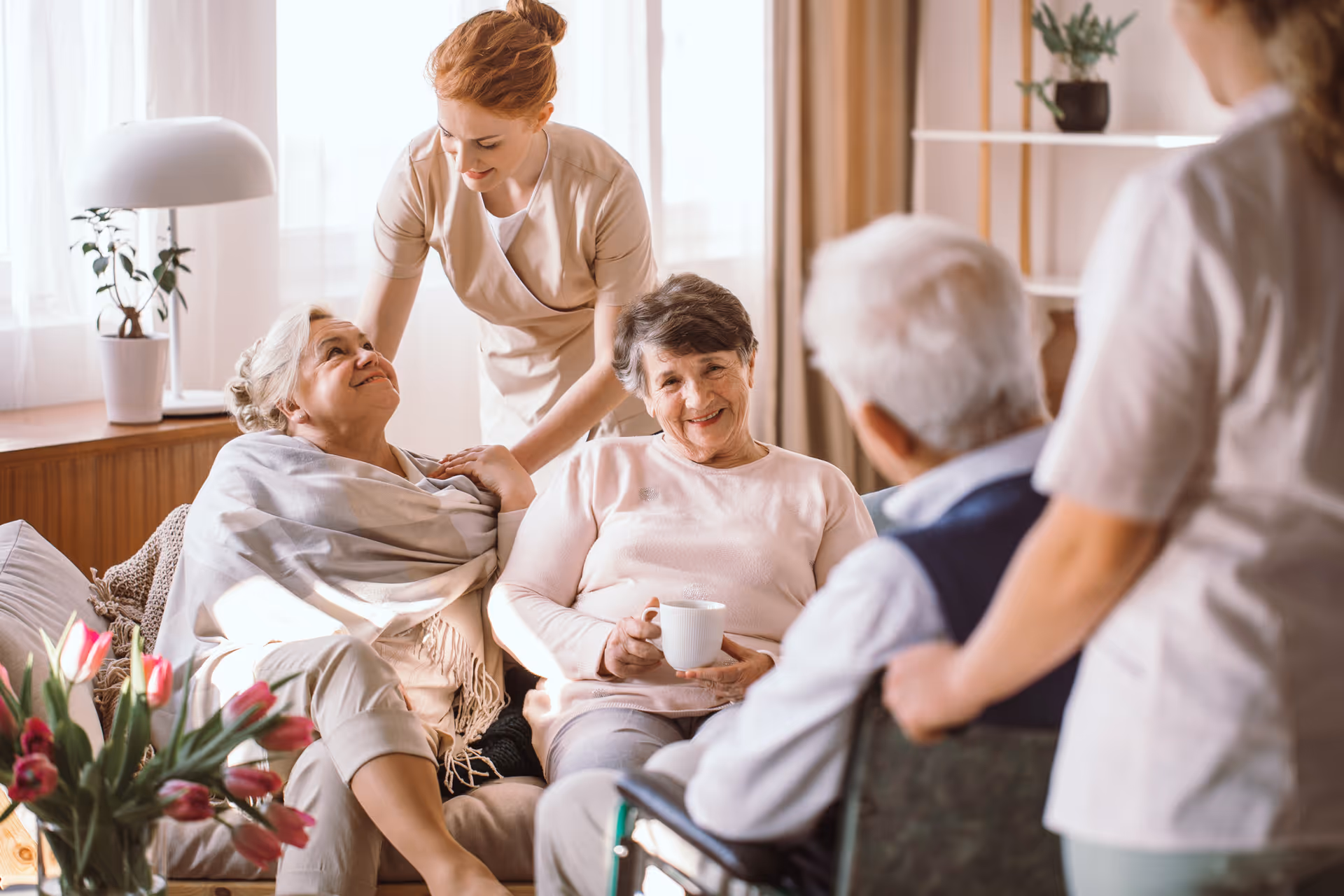 A group of elderly people sitting and interacting in a cozy living room with two caregivers attending to them. One elderly woman is wrapped in a blanket, another is holding a cup, and a man in a wheelchair is facing them. The room is warmly lit with a lamp, plants, and flowers on a table.