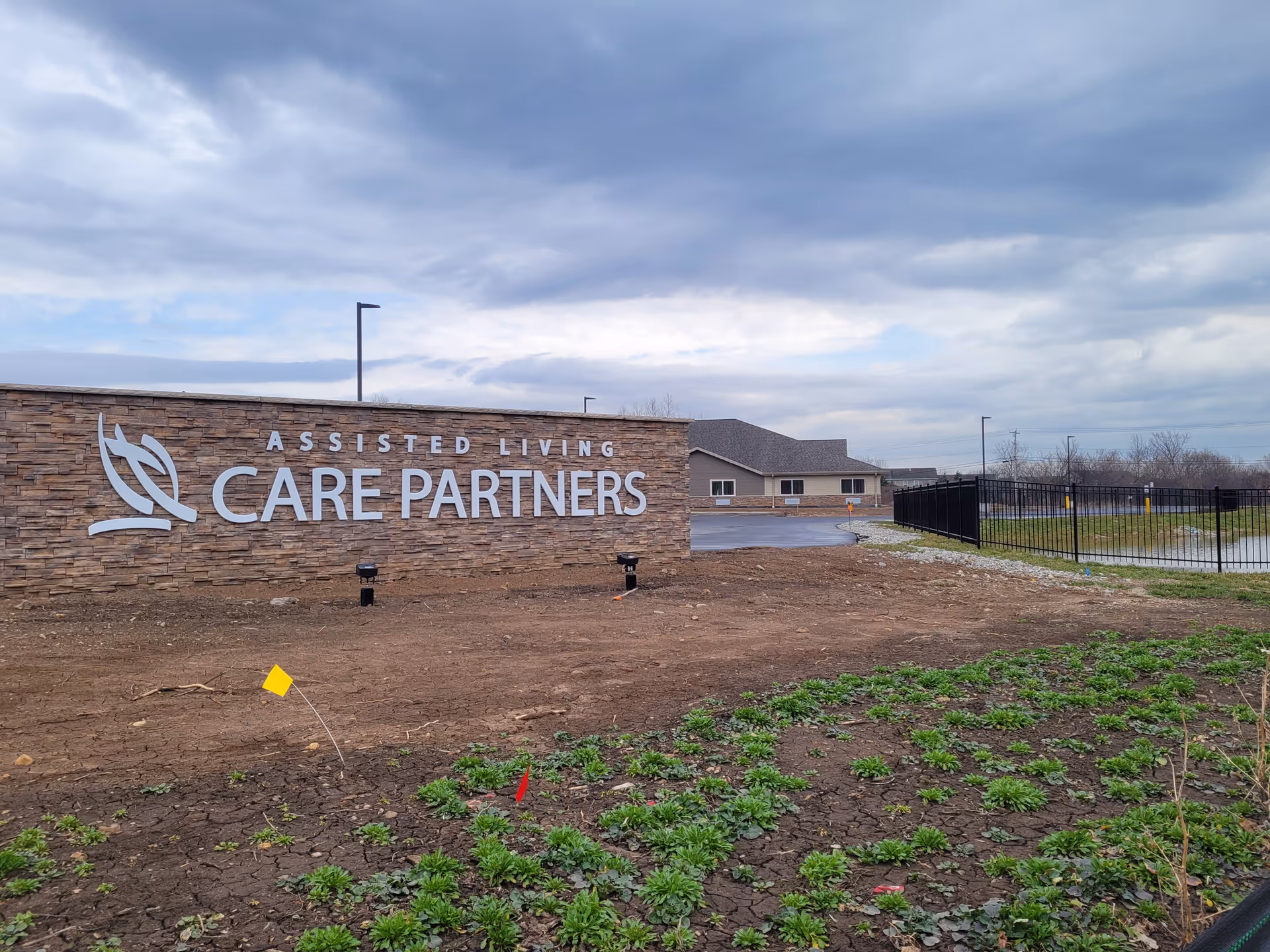 Outdoor view of the entrance sign for Care Partners Assisted Living, mounted on a stone wall with a cloudy sky overhead. The building and parking area are visible in the background, along with some greenery and a black metal fence.