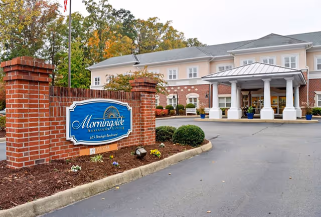 Exterior view of a senior living facility with a brick sign in the foreground that reads 'Morningside Assisted Living 655 Dunleigh Boulevard'. The building has a covered entrance with white columns and a driveway leading up to it. Trees with autumn foliage are visible in the background.