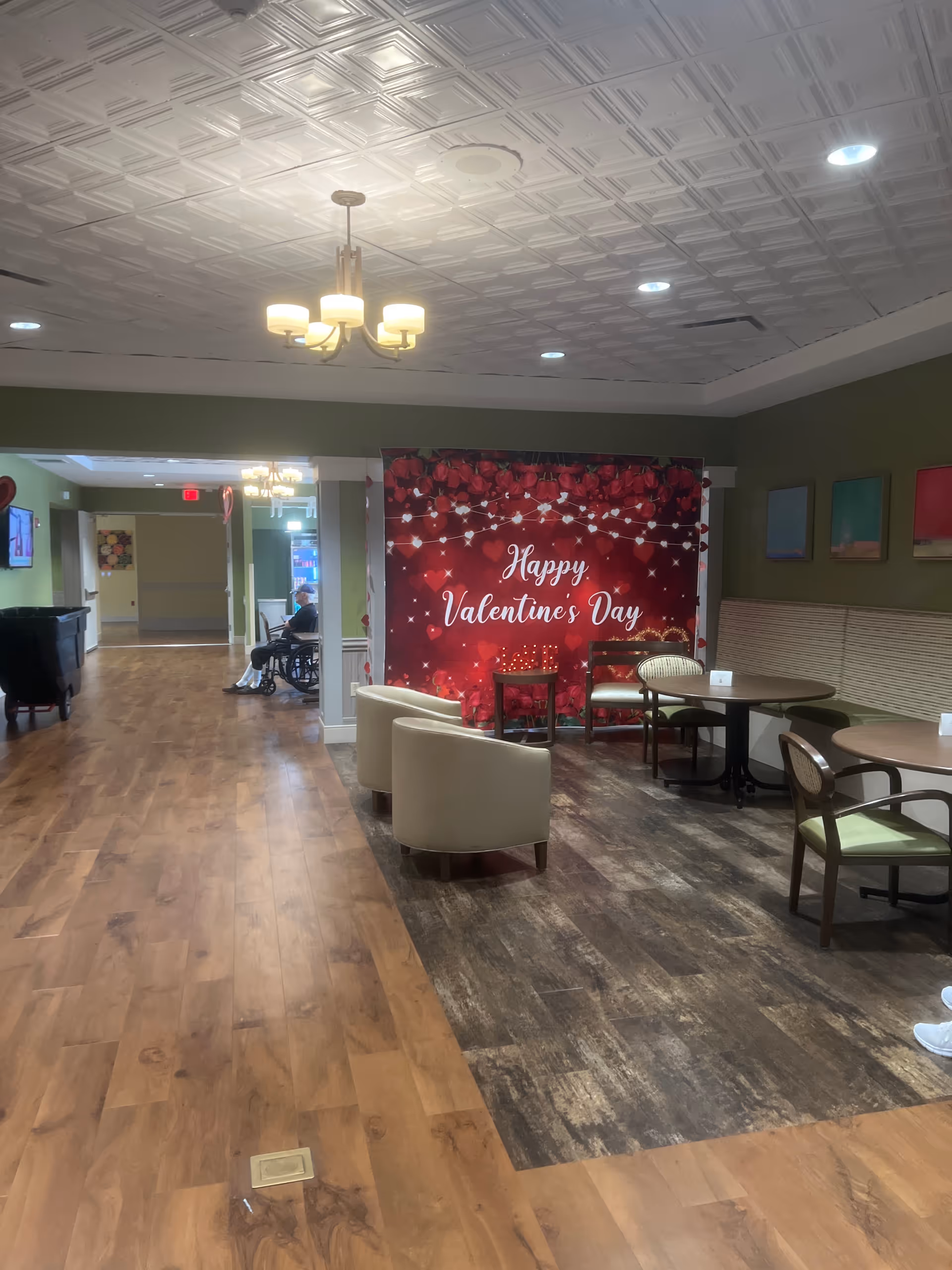Lounge area with a Valentine backdrop, tables, chairs, and a hallway in a senior living center.