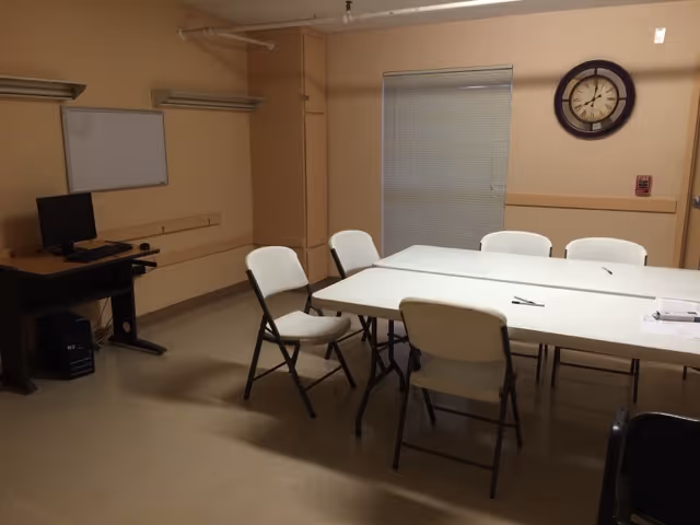 A small meeting or activity room with a rectangular table surrounded by six white folding chairs. There is a computer on a desk against the wall, a whiteboard mounted on the wall, a window with closed blinds, and a round wall clock above the window. The room has beige walls and a beige floor.