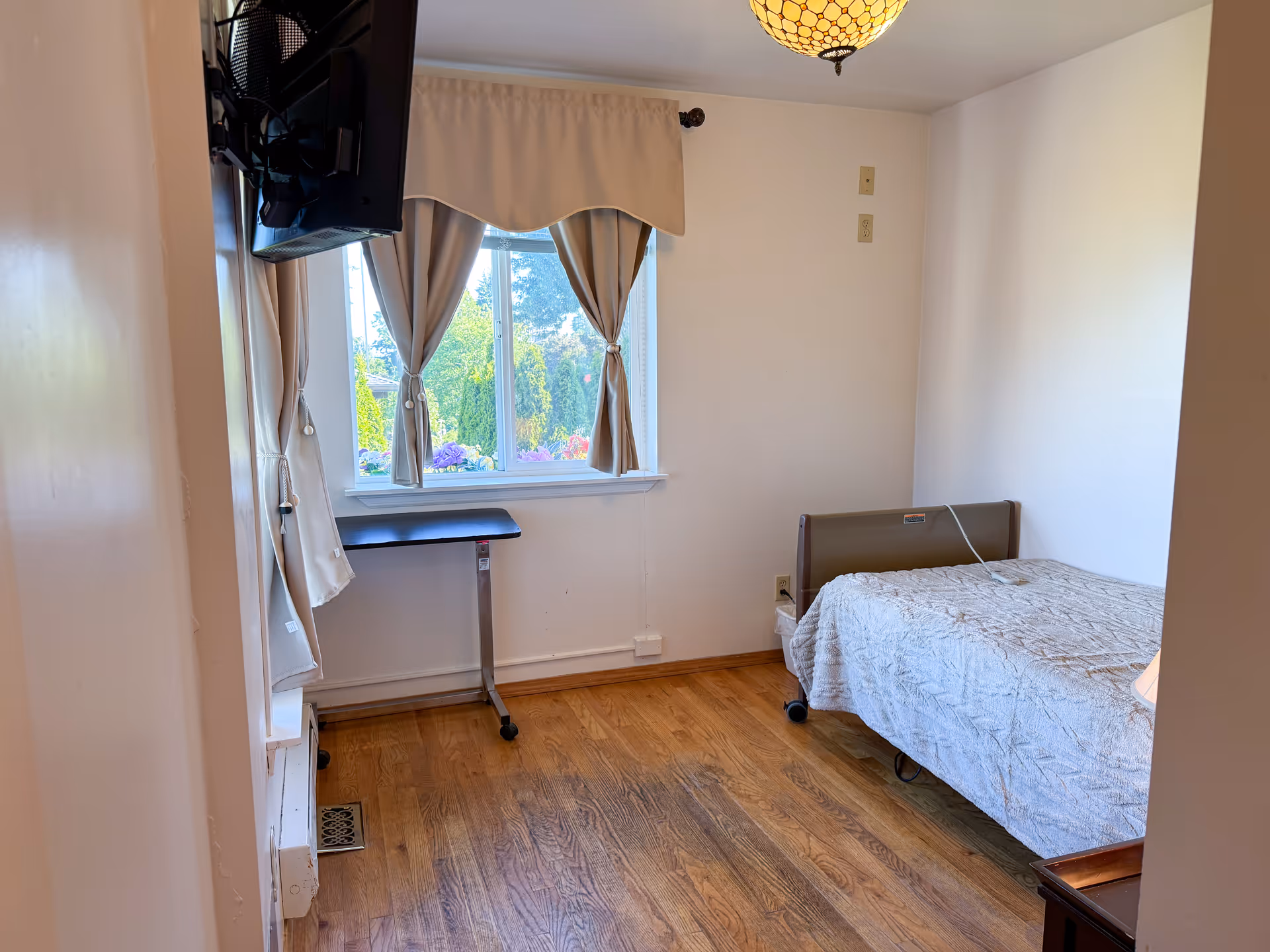 Small senior living bedroom with single bed, window, curtains, and a wall-mounted TV.
