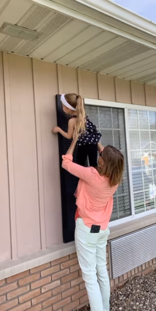 A woman in a pink shirt is lifting a young girl wearing a black outfit and white headband up to touch or look at a black shutter on the exterior wall of a building with beige siding and a window.