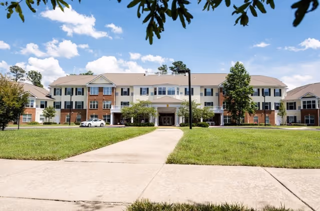 Front exterior view of a large, three-story senior living facility building with a mix of brick and light-colored siding. The building has multiple windows, a central entrance with a covered porch, and is surrounded by a well-maintained lawn and a concrete walkway leading to the entrance. A few trees and a parked white car are visible near the building under a partly cloudy blue sky.