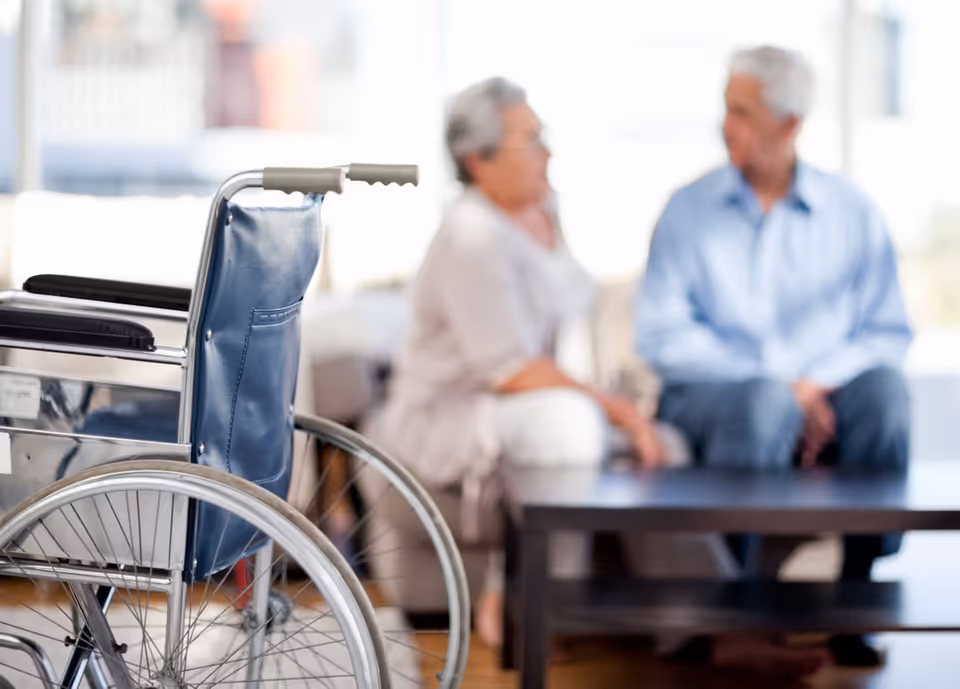 Wheelchair in foreground with blurred elderly couple seated in a living room.
