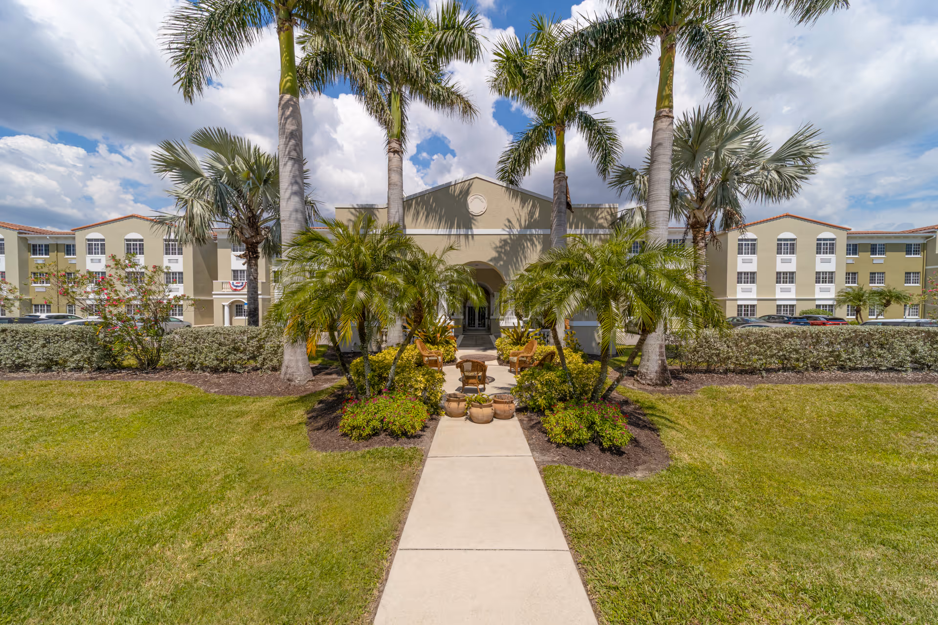 Front exterior view of DeSoto Palms Assisted Living Community building with a walkway leading to the entrance, surrounded by palm trees, shrubs, and a well-maintained lawn under a partly cloudy sky.