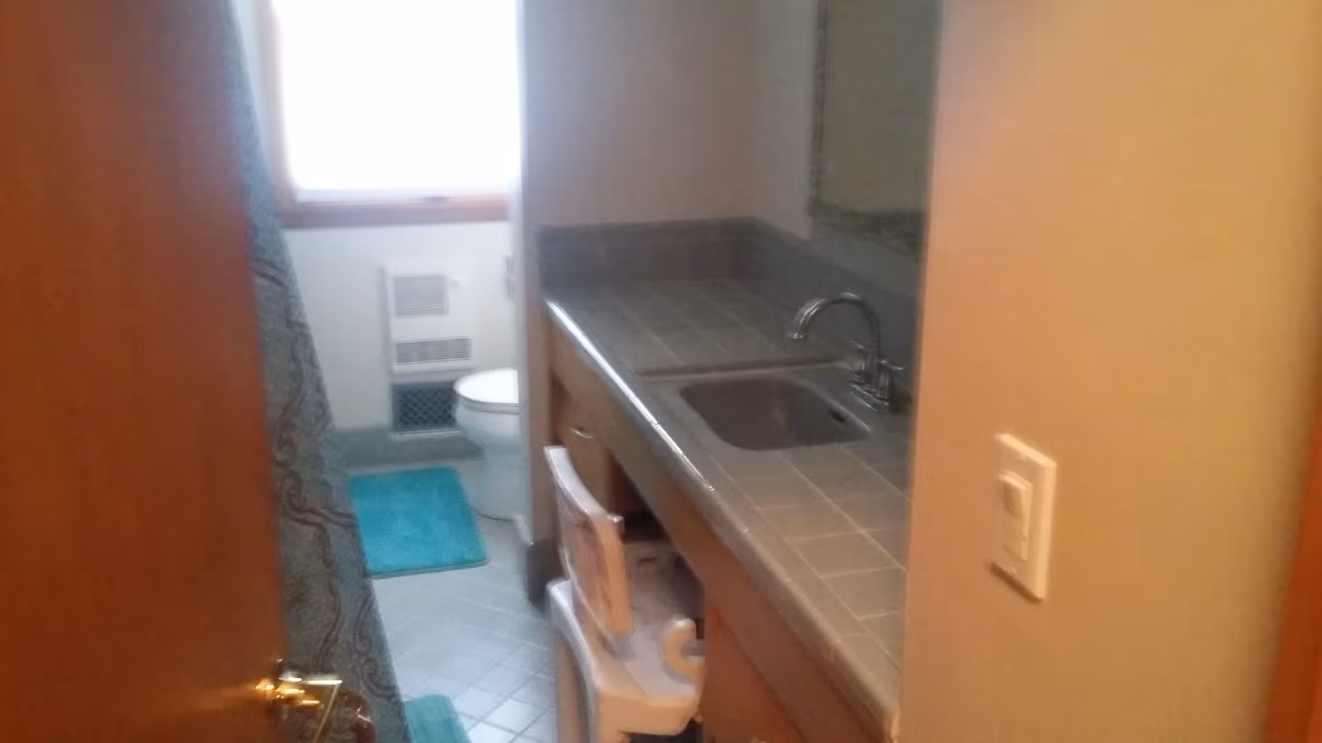 View into a small bathroom with a tiled countertop and sink on the right side. A toilet is visible in the background near a window with a blue bath mat on the floor. The bathroom door is partially open.