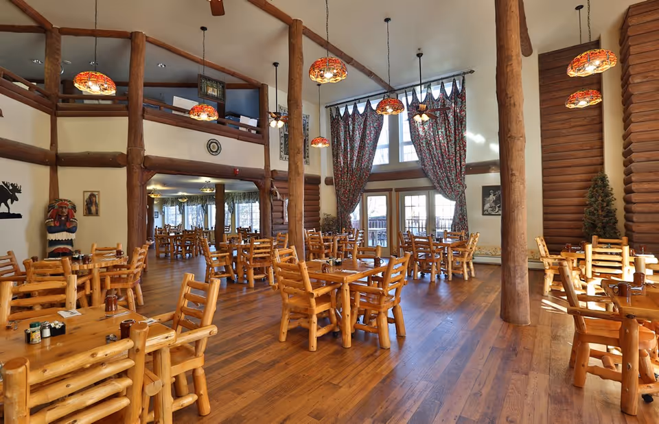 Spacious log-cabin style dining room with wooden tables and chairs, high ceilings, hanging stained-glass lamps, and large draped windows.