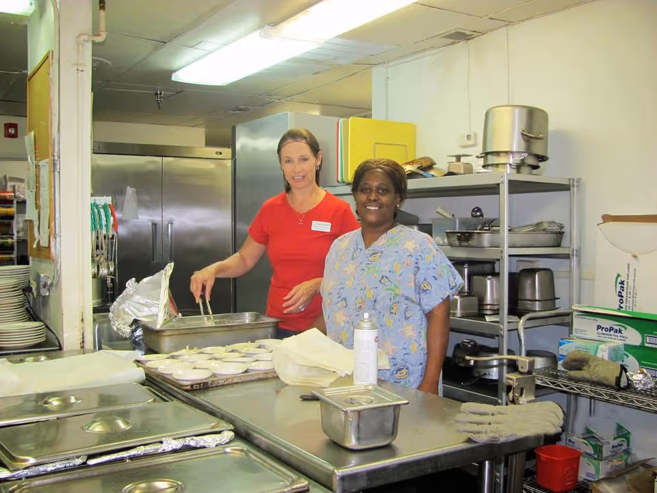 Two staff members smiling while preparing food in a commercial kitchen with stainless-steel counters and shelving.