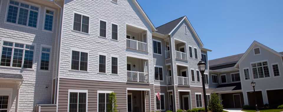 Exterior view of a multi-story residential building with white siding and multiple windows, balconies, and a few street lamps in front. The building has a modern design with a combination of horizontal and shingle siding and a well-maintained lawn area.