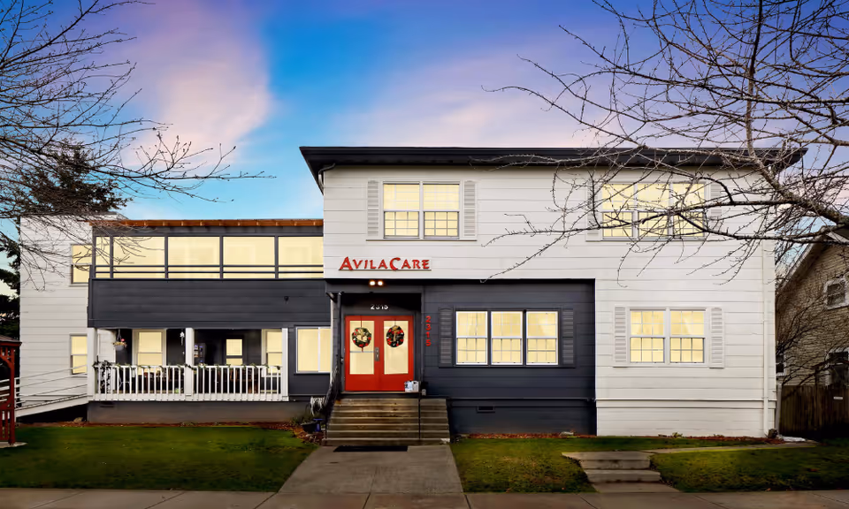Front exterior view of a two-story building named AvilaCare, with white and dark gray siding, red double doors decorated with wreaths, multiple windows, and a small porch area. The sky is clear with a gradient from blue to pink, and leafless trees frame the building.
