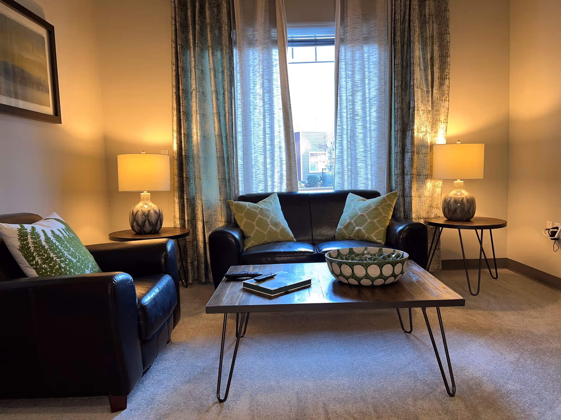 A cozy living room with a black leather loveseat and armchair, each adorned with green patterned pillows. A wooden coffee table with metal legs sits in front of the loveseat, holding a decorative bowl and two remote controls. Two matching table lamps with beige shades are placed on round wooden side tables on either side of the loveseat. Light filters through a window with sheer curtains behind the loveseat, and a framed picture hangs on the left wall.