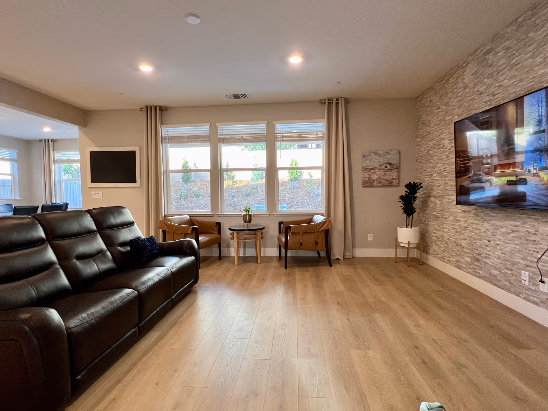 A spacious living room with a leather sofa, chairs, large windows, TV, and stone accent wall.