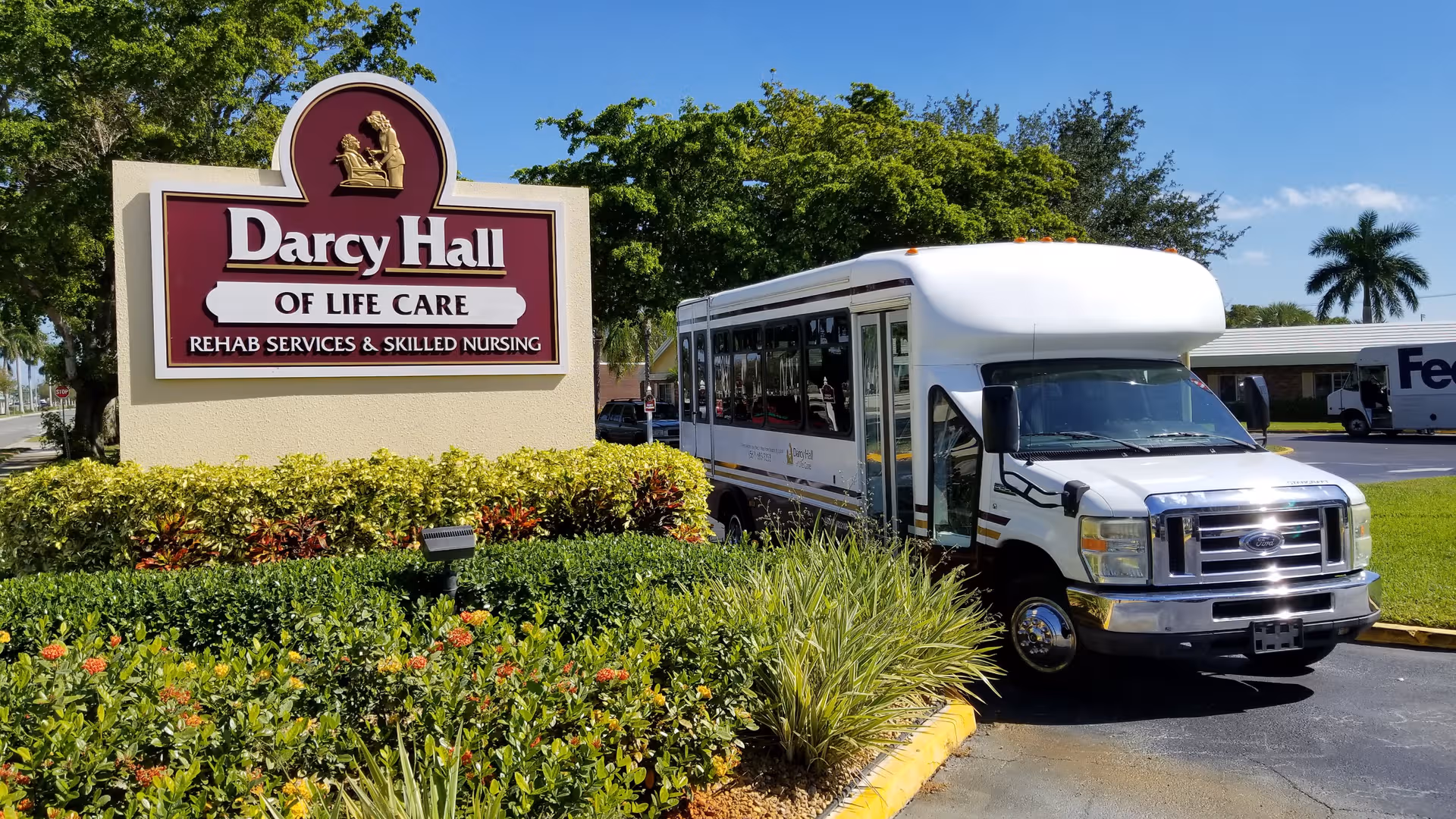 Exterior sign for Darcy Hall of Life Care with a shuttle bus parked beside landscaped grounds.