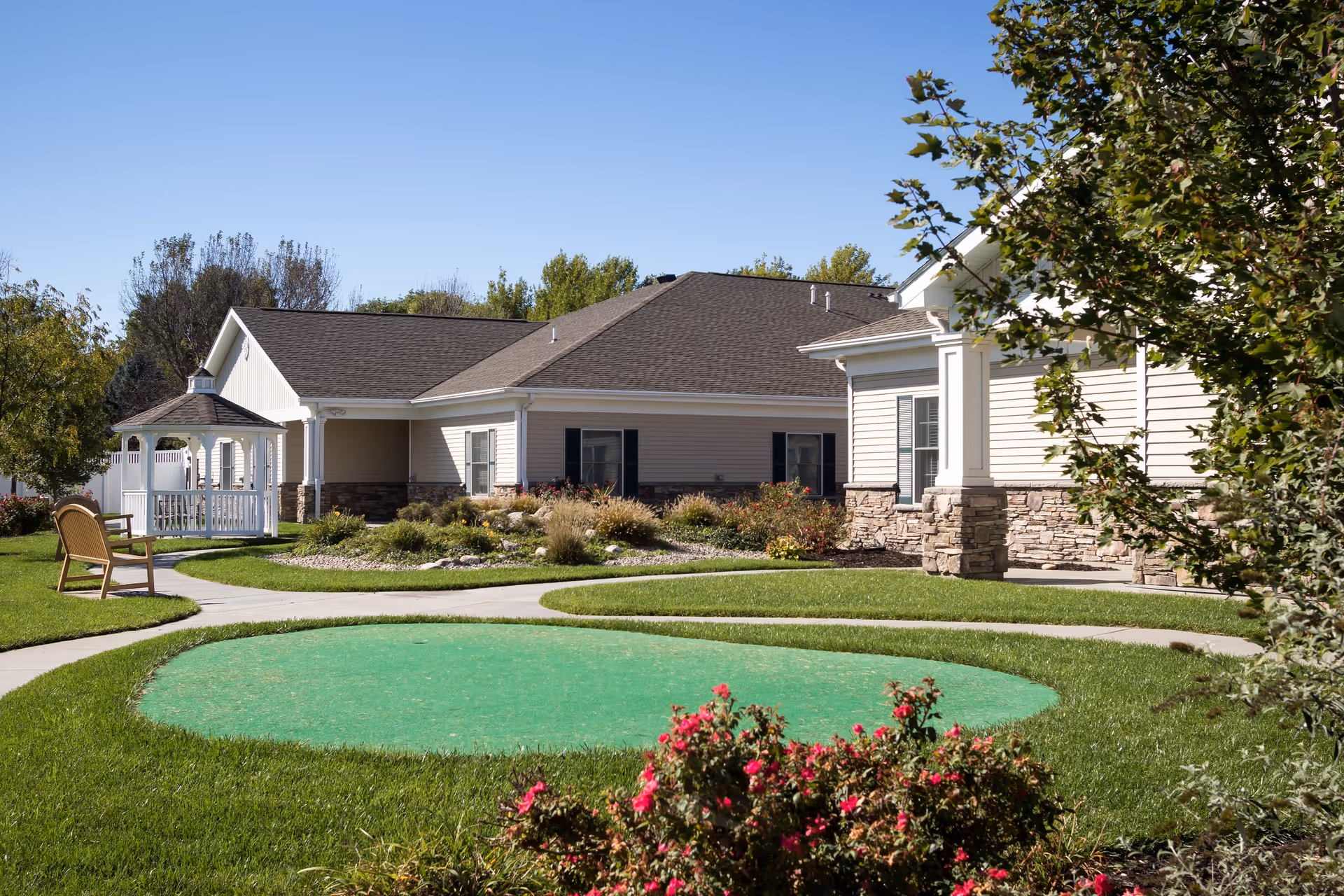 Outdoor view of a senior living facility with a well-maintained lawn, a small putting green, a wooden bench, a white gazebo, and beige buildings with stone accents under a clear blue sky.