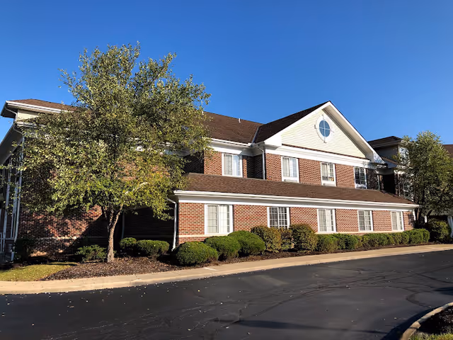 Exterior view of a two-story brick building with white trim and a brown roof, surrounded by neatly trimmed bushes and trees under a clear blue sky.