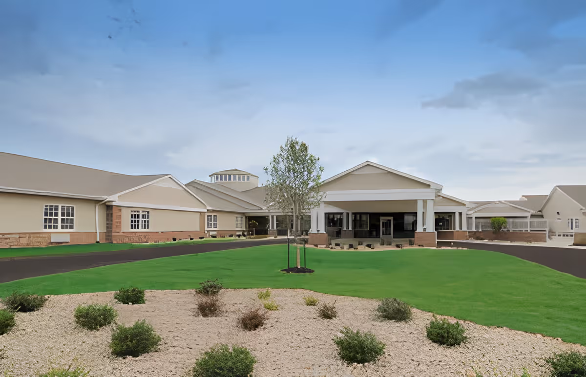 Exterior view of Aspen Place Health Campus showing a large single-story building with beige walls and a covered entrance supported by white columns. The foreground features a landscaped area with small bushes and a young tree, surrounded by green grass and a paved driveway.