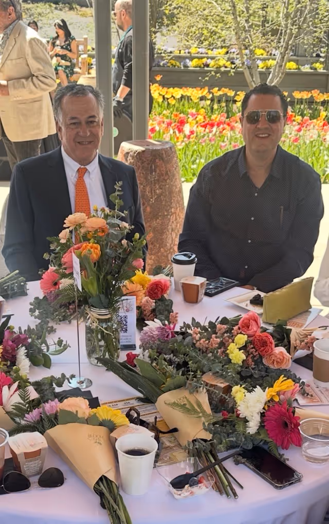 Two men in smart casual attire seated at a flower-adorned outdoor table.