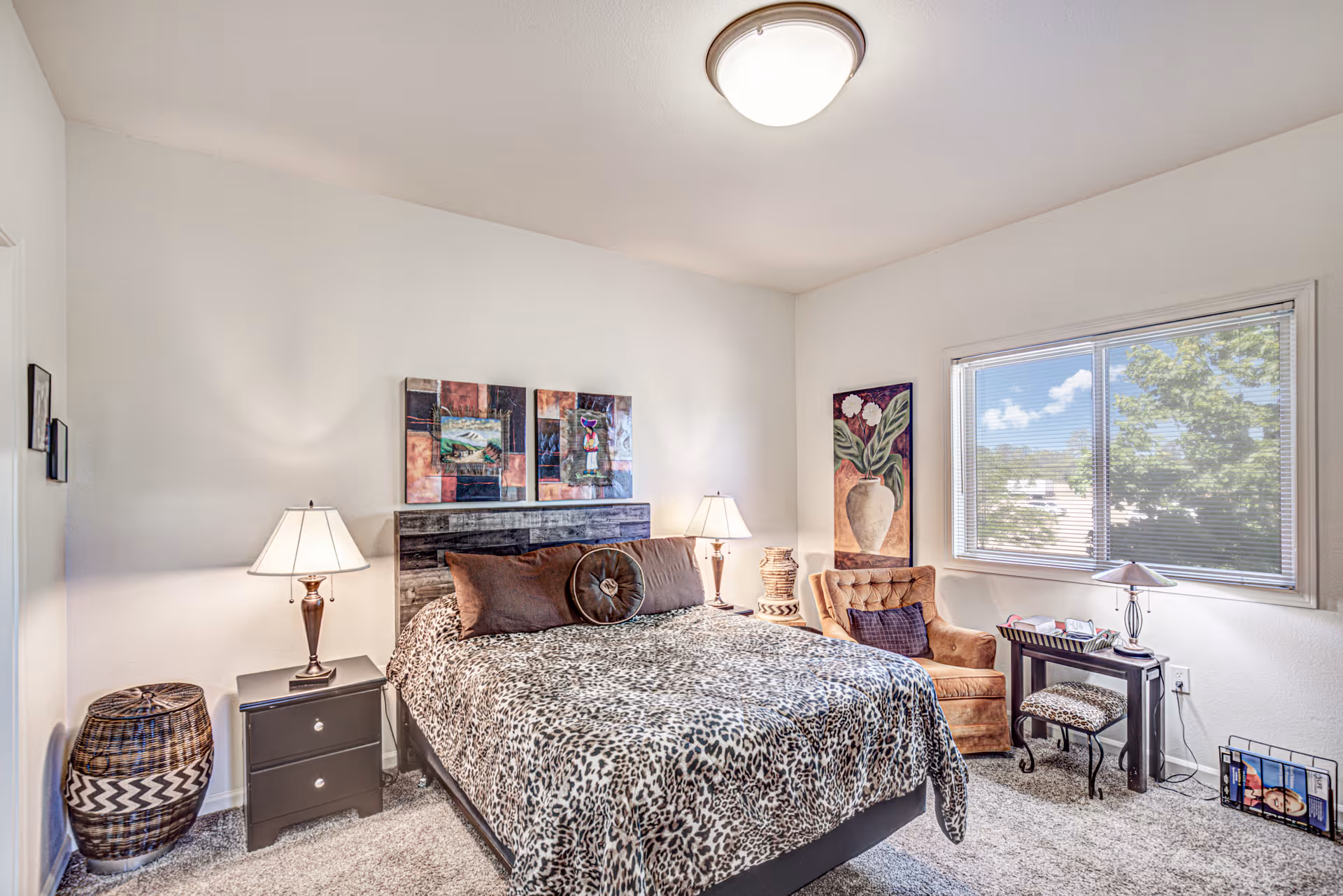 A cozy bedroom with a large bed covered in a leopard print blanket, two bedside tables each with a lamp, a comfortable armchair with a pillow, a small desk with a lamp and a stool, and a window showing a view of trees outside. The walls are decorated with colorful paintings.