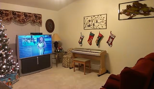 Cozy living room with a lit Christmas tree, a TV showing a presenter, an electronic keyboard with three hanging stockings, and red upholstered seating.