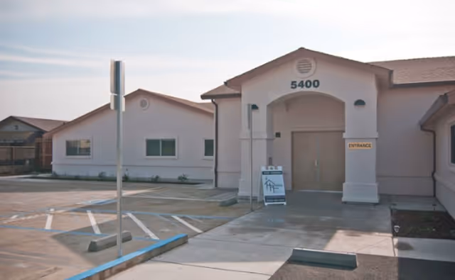 Exterior view of Hospitality House Assisted Living & Memory Care building with the address number 5400 above the entrance. The building is single-story with a light-colored facade and a parking area in front, including a handicapped parking space and a sign near the entrance.