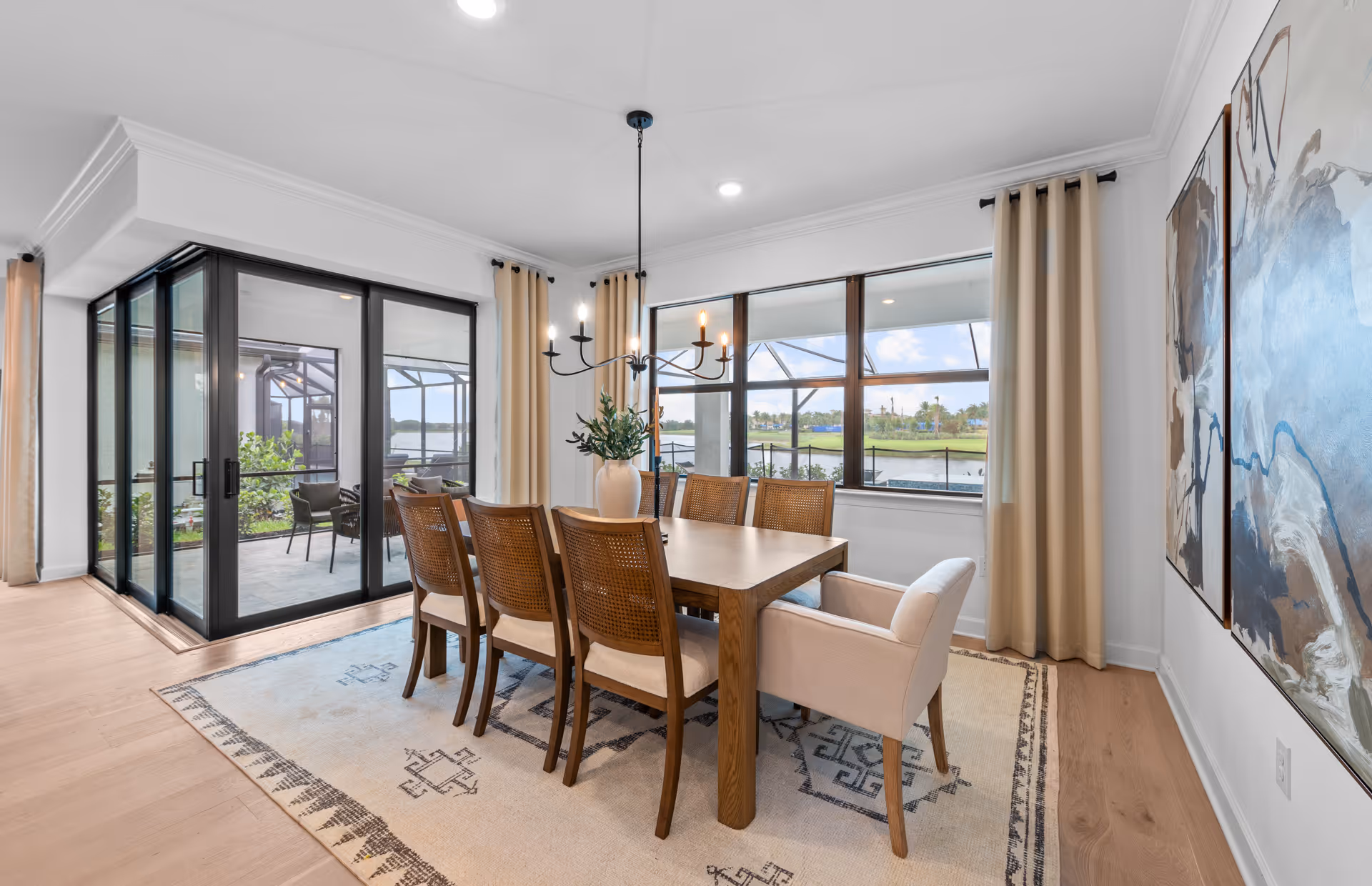 Bright dining room with a wooden table surrounded by six chairs, including one armchair. Large windows with beige curtains provide a view of a screened patio and a scenic outdoor landscape with water and greenery. Two abstract paintings hang on the wall, and a modern chandelier hangs above the table.