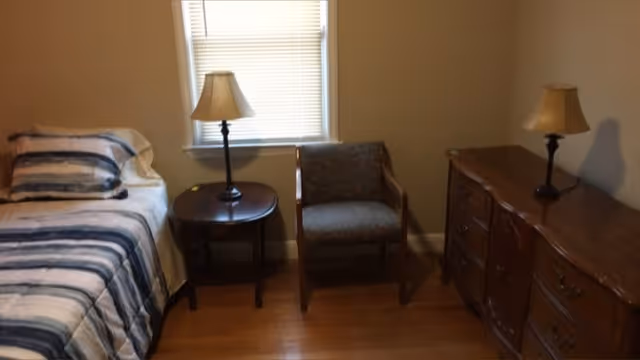 Small furnished bedroom with a single bed, nightstand and lamp, an armchair, and a wooden dresser beneath a window.