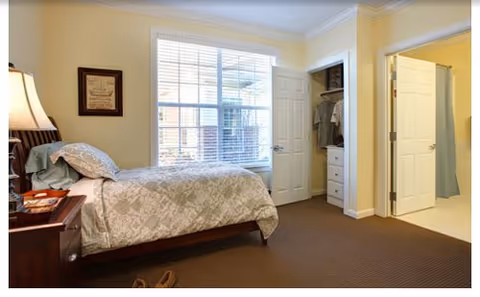 Bright bedroom with bed, nightstand, lamp, window blinds, closet, and doorway to hall.