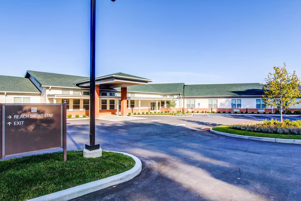 Front entrance and driveway of a single-story senior living building with a covered drop-off and a sign pointing to short-stay and exit.