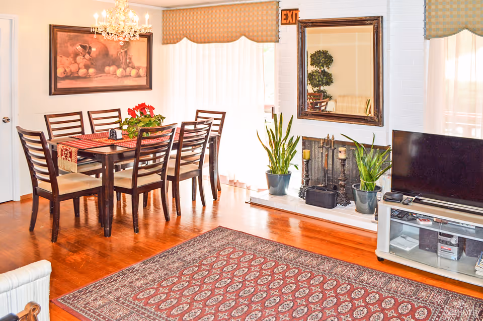 A bright dining and living area with a wooden dining table and six chairs, a chandelier above the table, a large framed painting on the wall, and a window with sheer curtains. The living area features a fireplace with plants on either side, a large mirror above the fireplace, a TV on a stand, and a patterned rug on the wooden floor.