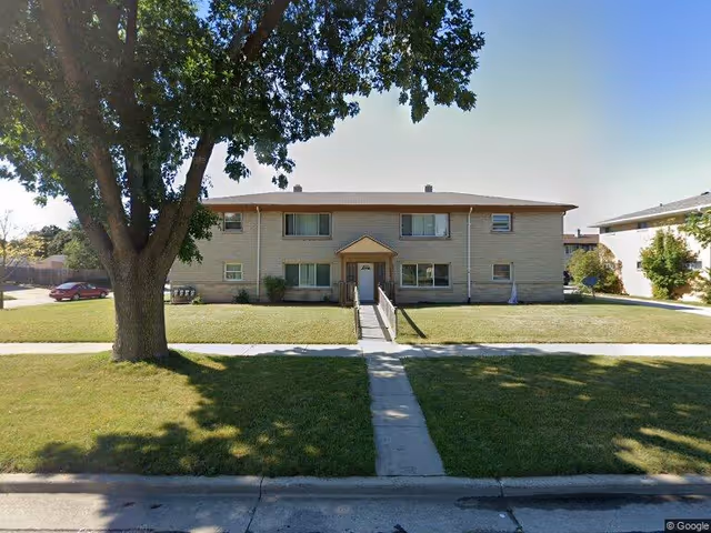 Two-story brick apartment building with a central entrance, front lawn, sidewalk, and a large tree in the foreground.