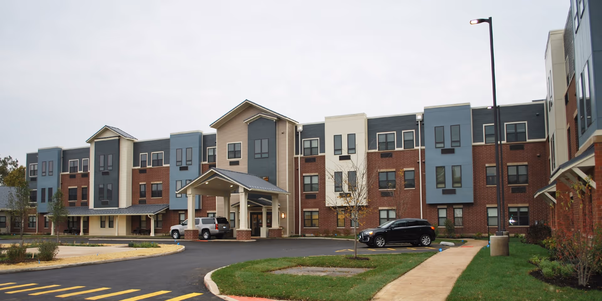 Exterior view of a modern three-story senior living facility with a covered entrance, multiple windows, a driveway with parked cars, and landscaped green areas.