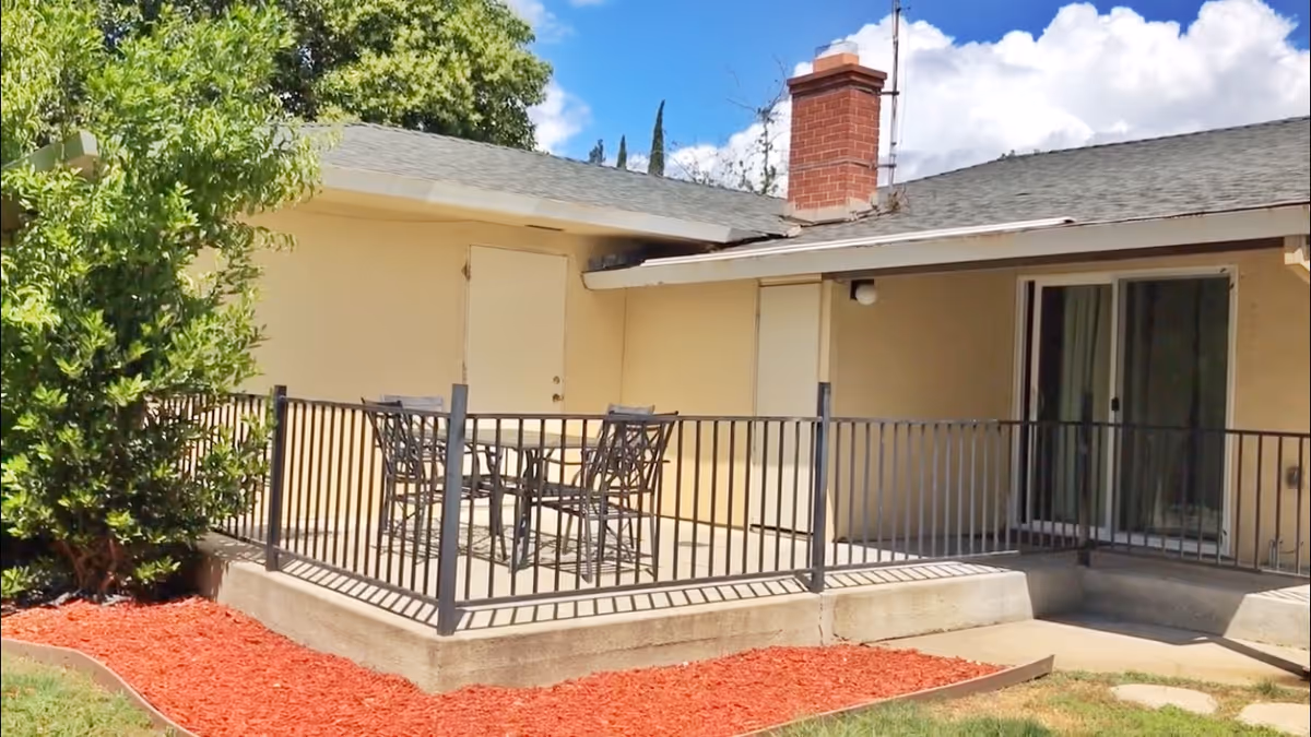 Outdoor patio area with a metal table and four chairs surrounded by a black metal railing. The patio is attached to a beige building with a chimney and sliding glass doors. There is a tree and red mulch landscaping around the patio.