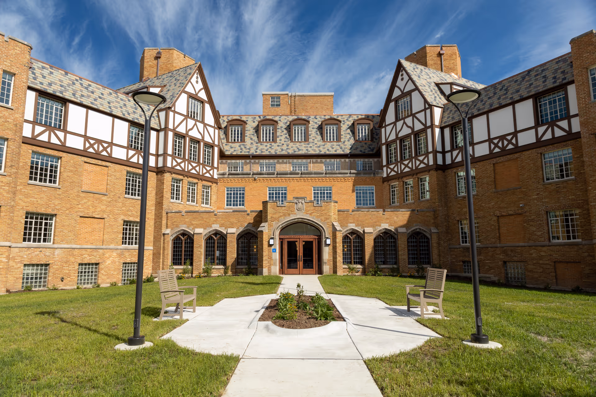 Brick Tudor-style building with a central arched entrance, courtyard path, benches and lamp posts under a blue sky.
